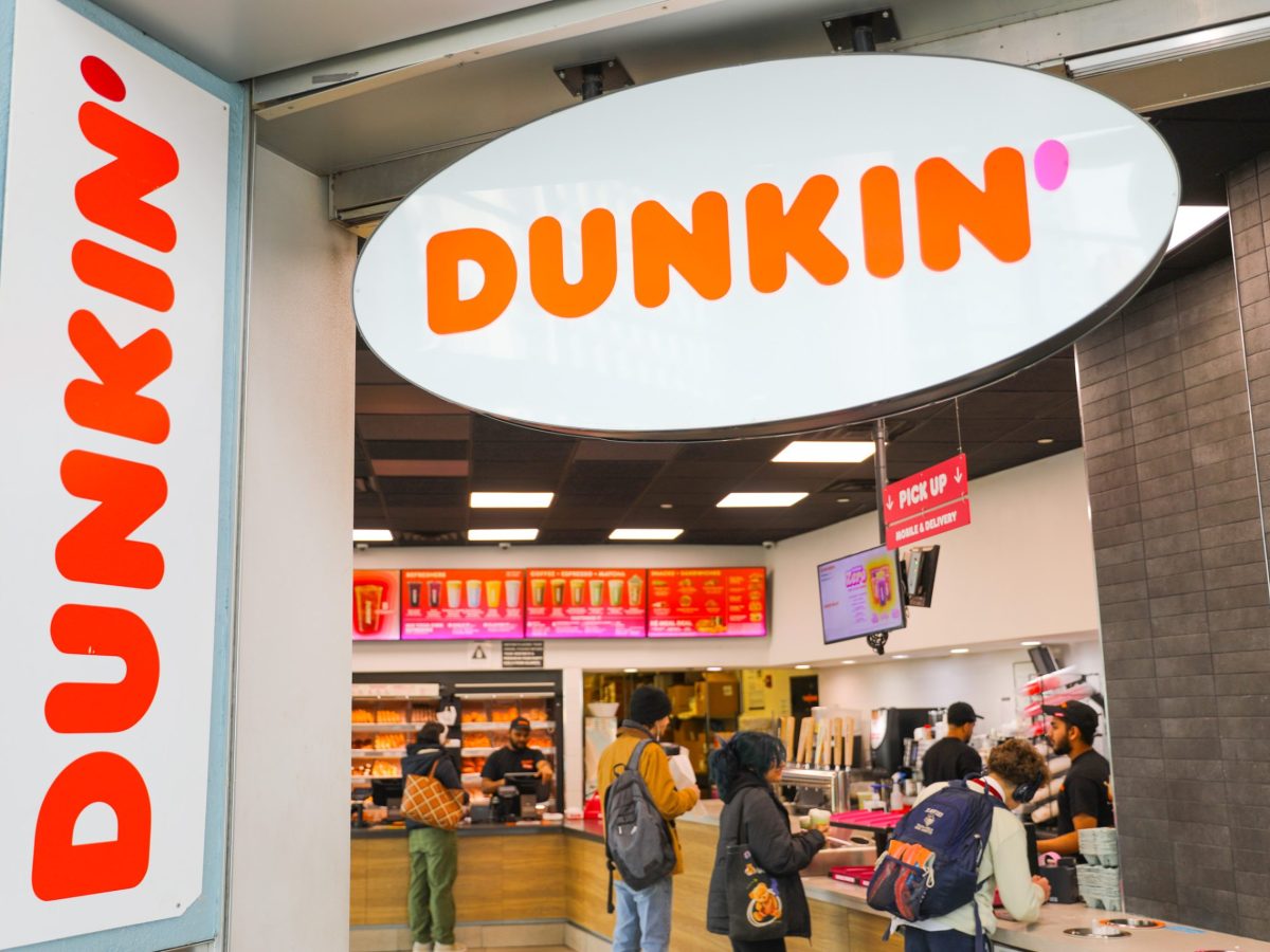 The Dunkin sign in bright orange is on the top and left side of the frame. Four customers are being helped at the counter by Dunkin employees wearing black clothes with the Dunkin Donuts icon.