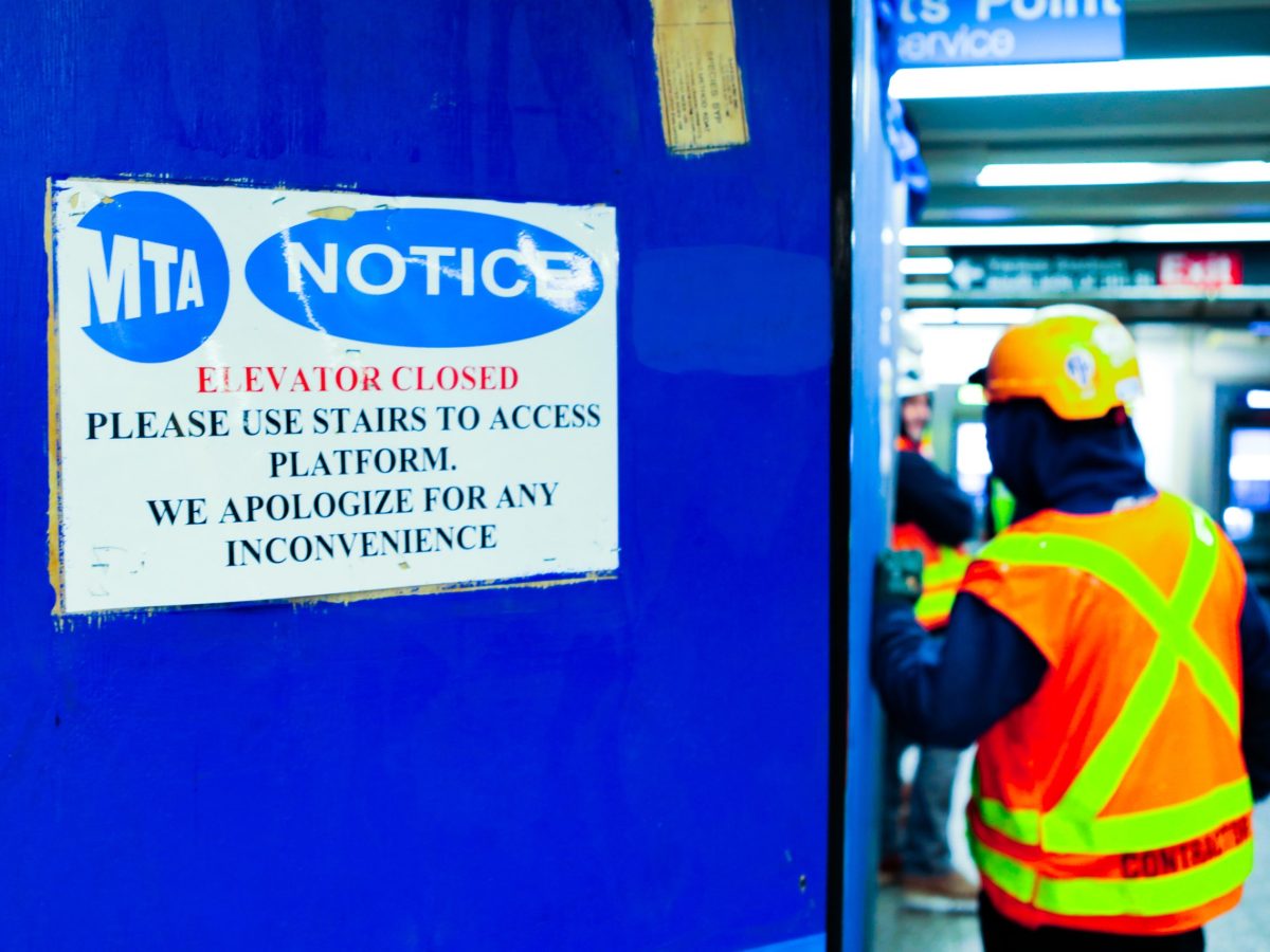 A laminated MTA sign on a blue construction site wall reads "Elevator closed, Please use stairs to access platform. we apologize for an inconvenience. A construction worker in a orange safety vest and hard hat stand out of focus to the right of the site.