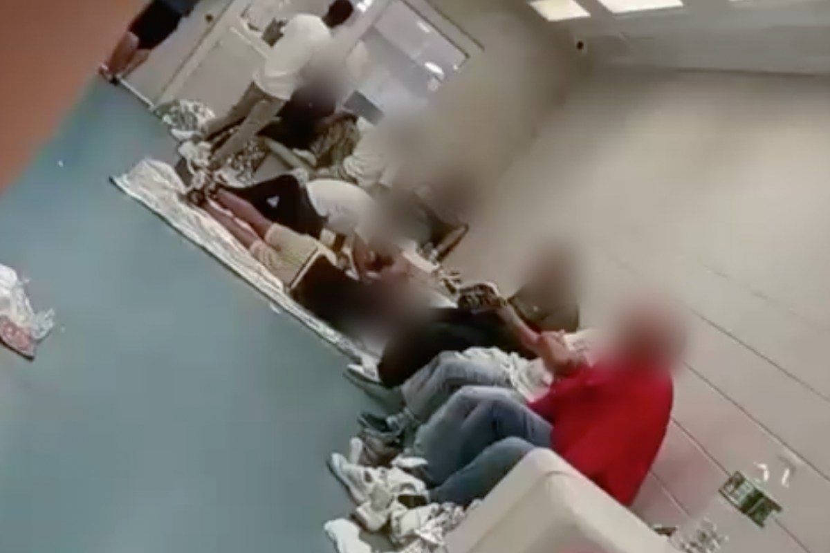 A dozen people with their faces blurred sit and lay side by side on a concrete floor. A door with a glass window can be seen on the far side. Multiple people on the floor lay on thin emergency blankets.
