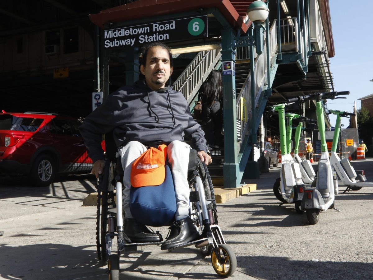 Rodolfo wears a black hoodie and grey sweatpants. With both hands on his wheelchair he looks straight ahead with the middletown road subway behind him.