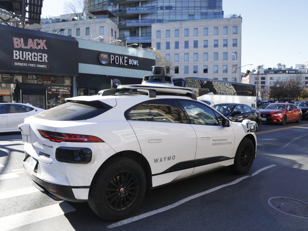 Waymo vehicle on canal street. The white vehicle says autonomous mode on the side. Black cameras that can be seen on the top and sides. Black burger and POKE bowl restaurant signs are behind the self driving car.