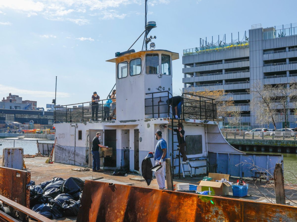 A white boat with visible rust sits in Anable boat basin off the east river. Seven people can be seen cleaning with trash bags and supplies on the deck. Buildings can be seen behind the boat across the water.