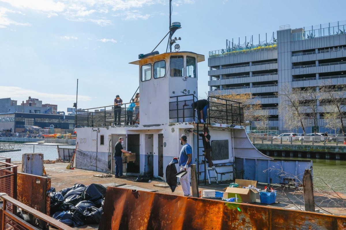 A white boat with visible rust sits in Anable boat basin off the east river. Seven people can be seen cleaning with trash bags and supplies on the deck. Buildings can be seen behind the boat across the water.