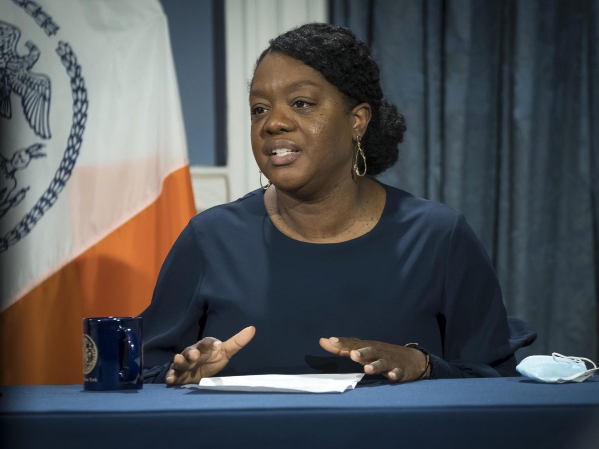 Sideya Sherman wears a blue blouse and gold earrings as she speaks from a panel with the city council flag behind her